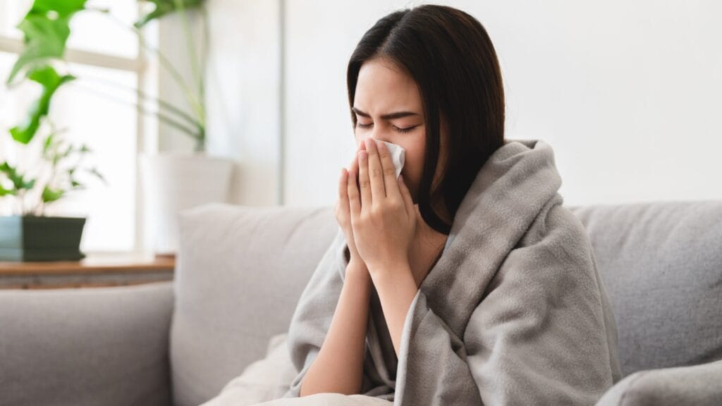 Woman with post nasal drip symptoms blowing her nose, wrapped in a blanket.